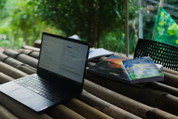 Un ordinateur portable posé sur une table en bambou, entouré de livres et de documents, dans un environnement verdoyant et luxuriant, créant une atmosphère de travail paisible et productive.