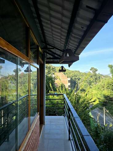 Un balcon moderne aux balustrades noires surplombe des arbres verdoyants et des collines lointaines. Des fenêtres en verre reflètent le paysage et des lampes suspendues sont accrochées au toit en pente.