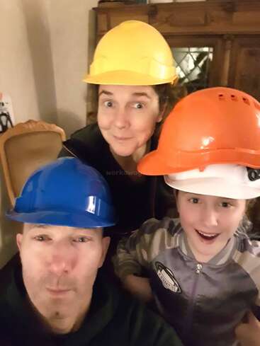 L'image montre une famille de trois personnes portant des casques de protection, posant pour un selfie, avec le père à gauche, la mère derrière et l'enfant à droite, tous souriants.