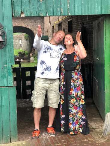 La imagen representa a un hombre y una mujer de pie, juntos, con los brazos en alto en un gesto de celebración o saludo, delante de un rústico edificio de ladrillo.