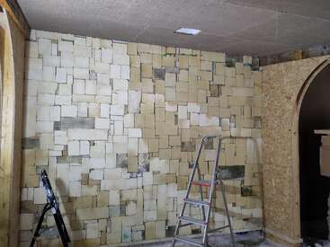 Esta imagen muestra una pared interior sin terminar, revestida con baldosas rectangulares de piedra. Se ven dos escaleras, materiales de construcción y una puerta con arco, lo que indica que se están llevando a cabo obras de renovación.