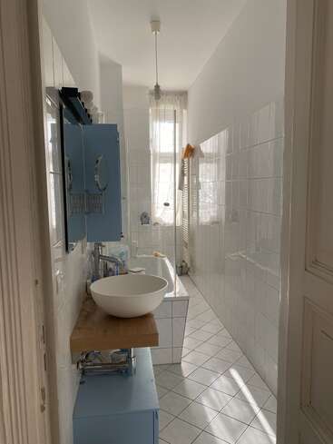 Das ist ein schmales, weiß gefliestes Badezimmer mit einem runden Waschbecken auf einem Holzwaschtisch, blauen Schränken, einer Badewanne, einem Wandspiegel und Tageslicht durch ein Fenster.