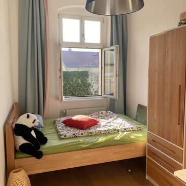 Une petite chambre confortable avec un lit en bois, une literie verte, un oreiller rouge, une peluche panda, une armoire, une fenêtre ouverte, des rideaux bleus et la lumière du soleil qui pénètre à l'intérieur.