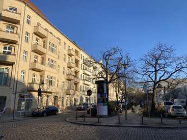 Das Bild zeigt eine sonnige Straße in der Stadt mit gelben Wohnhäusern, geparkten Autos, kahlen Bäumen, einer runden Werbesäule, Kopfsteinpflaster und einem strahlend blauen Himmel.