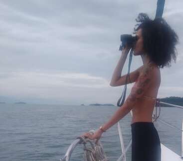 A woman stands on a boat, looking through binoculars at the horizon. She wears a bikini top and has curly hair, surrounded by calm water.