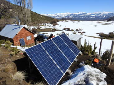 Eine rustikale Holzhütte steht inmitten einer verschneiten Landschaft und wird von Solarzellen betrieben. Die Berge und der blaue Himmel schaffen eine ruhige, abgelegene und nachhaltige Winterlandschaft.