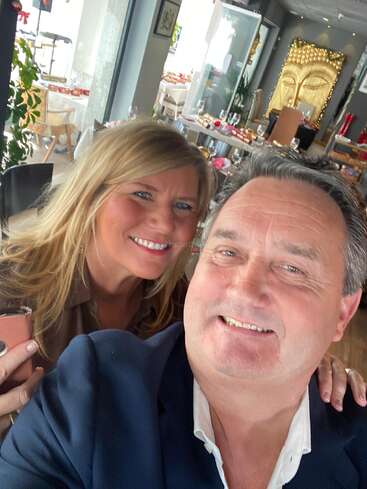 Ein lächelnder Mann und eine lächelnde Frau machen ein Selfie in einem schicken Restaurant mit eleganter Tischdeko und einem großen goldenen Buddha-Gesicht als Deko im Hintergrund.