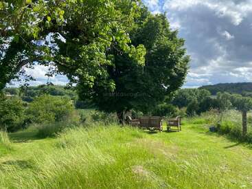 A peaceful grassy field with leafy trees, outdoor furniture arranged under shade, overlooking rolling green hills under a partly cloudy sky, creating a serene countryside atmosphere.