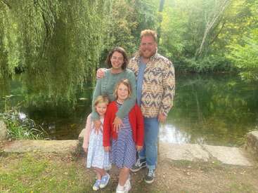 A happy family of four poses by a serene pond, surrounded by lush greenery and trees, enjoying a peaceful day outdoors together in nature.