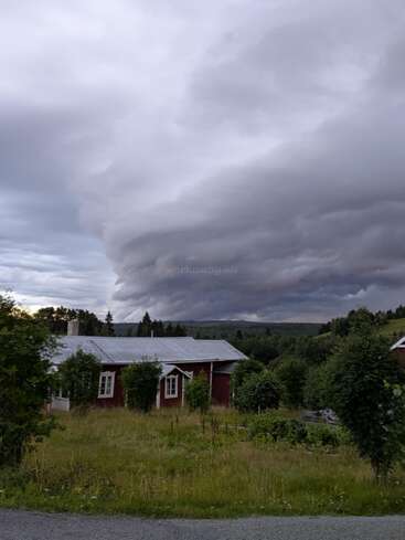 Una dramática y arremolinada nube de tormenta se cierne sobre una pequeña y rústica casa roja rodeada de árboles verdes y césped, situada en un apacible paisaje rural.