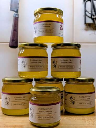 Six jars of honey labeled “Trolltjärns Gård Årets Honung” are neatly stacked on a wooden countertop, with kitchen utensils visible in the background.