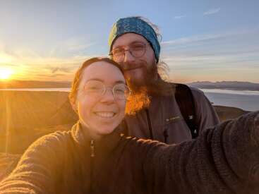 A smiling couple takes a selfie outdoors at sunset, bathed in golden light, wearing glasses and warm clothing with scenic hills and water in the background.