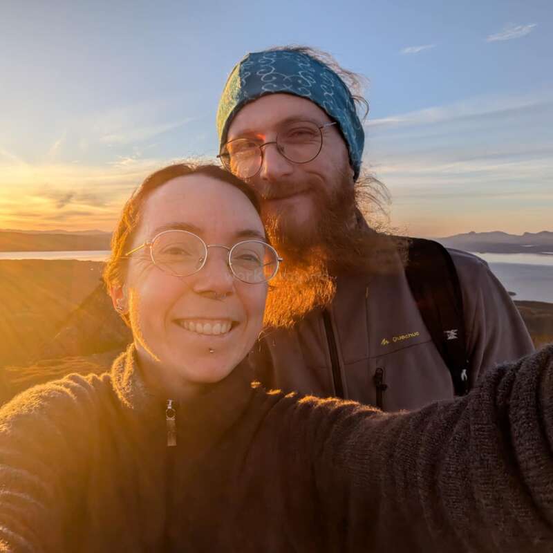 Una pareja sonriente se toma un selfie al aire libre al atardecer, bañada por una luz dorada, con gafas y ropa de abrigo, con unas pintorescas colinas y agua de fondo.