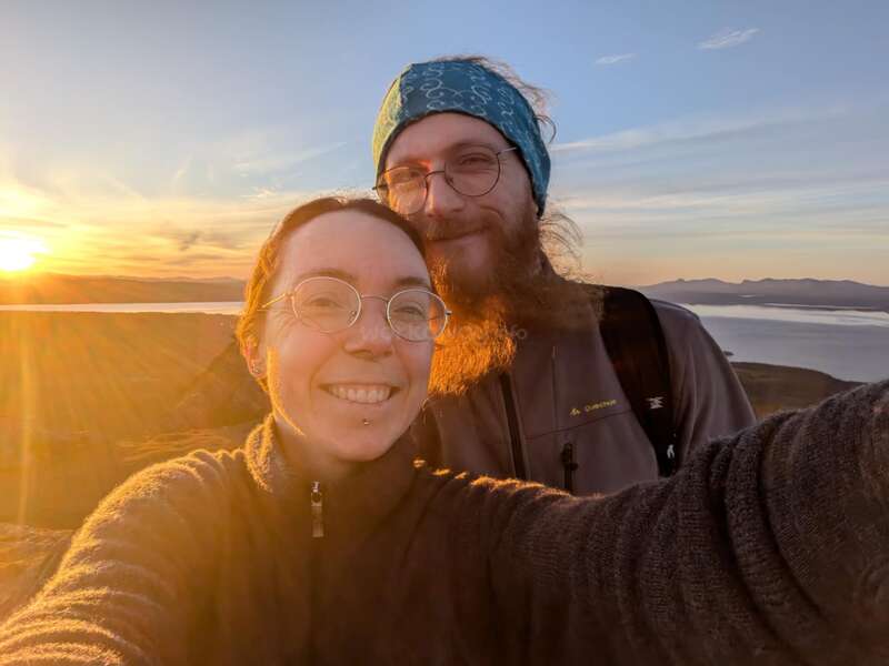 Un couple souriant prend un selfie en plein air au coucher du soleil, baigné dans une lumière dorée, portant des lunettes et des vêtements chauds, avec des collines pittoresques et de l'eau en arrière-plan.