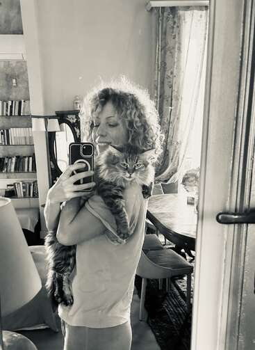 Uma mulher de cabelos cacheados tira uma selfie no espelho, sorrindo enquanto segura um gato fofo. O cômodo aconchegante tem estantes de livros, uma mesa de jantar e cortinas florais.