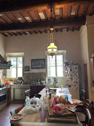 Uma cozinha rústica com vigas de madeira expostas, bancadas desorganizadas, luminária vintage pendurada, duas janelas claras, geladeira com ímãs e vários itens de cozinha espalhados pelo cômodo.