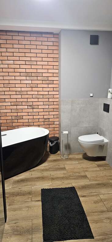 Este moderno cuarto de baño cuenta con una bañera exenta negra, inodoro flotante, paredes de ladrillo y gris, baldosas de madera, alfombra negra, papelera y accesorios mínimos.