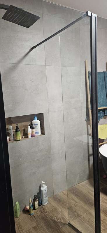 Una moderna ducha de baño con un gran rociador cuadrado, paredes de azulejos grises, mampara de cristal, suelo de madera, estante con artículos de aseo y toallas colgadas en una escalera de madera.