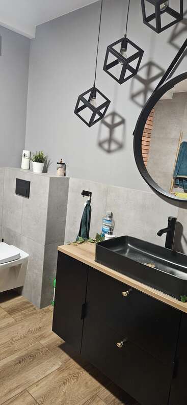 Un baño moderno con luces geométricas colgantes, un gran espejo redondo, lavabo negro, encimera de madera, paredes grises, plantas en macetas, toalla y una decoración ordenada.