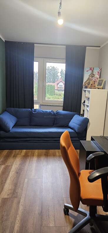 Una acogedora oficina en casa con suelo de madera, un sofá azul marino, una silla de oficina naranja, un escritorio negro, una estantería blanca, una ventana con cortinas negras y vistas al exterior.