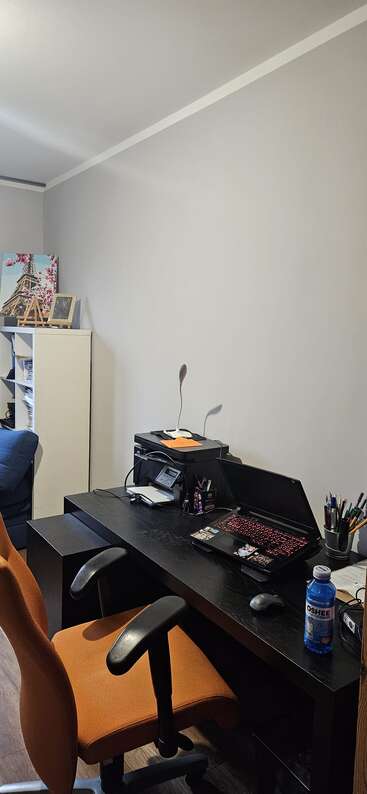 Un espacio de trabajo moderno con una silla naranja, escritorio negro, portátil abierto, impresora, teléfono, bolígrafos, botella de bebida Oshee, fotos y arte mural de la Torre Eiffel.