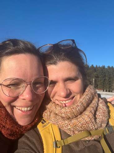 Two women smile warmly for a selfie outdoors on a sunny day, bundled in scarves. One wears glasses, while the other has glasses on her head.