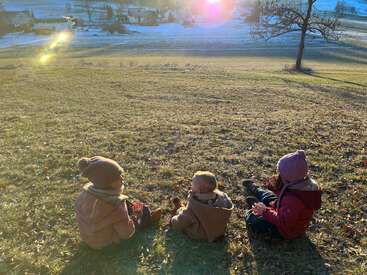 Three children in winter clothing sit on a grassy hill, enjoying the sunlight. Snow and houses are visible in the background, creating a peaceful, rural scene.