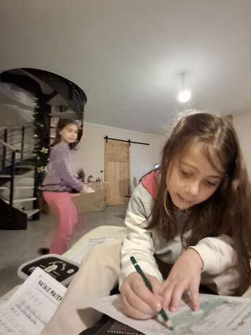 Una niña pinta con atención en un papel con un lápiz en primer plano, mientras otra niña se mueve cerca de una escalera de caracol en una acogedora sala de estar.