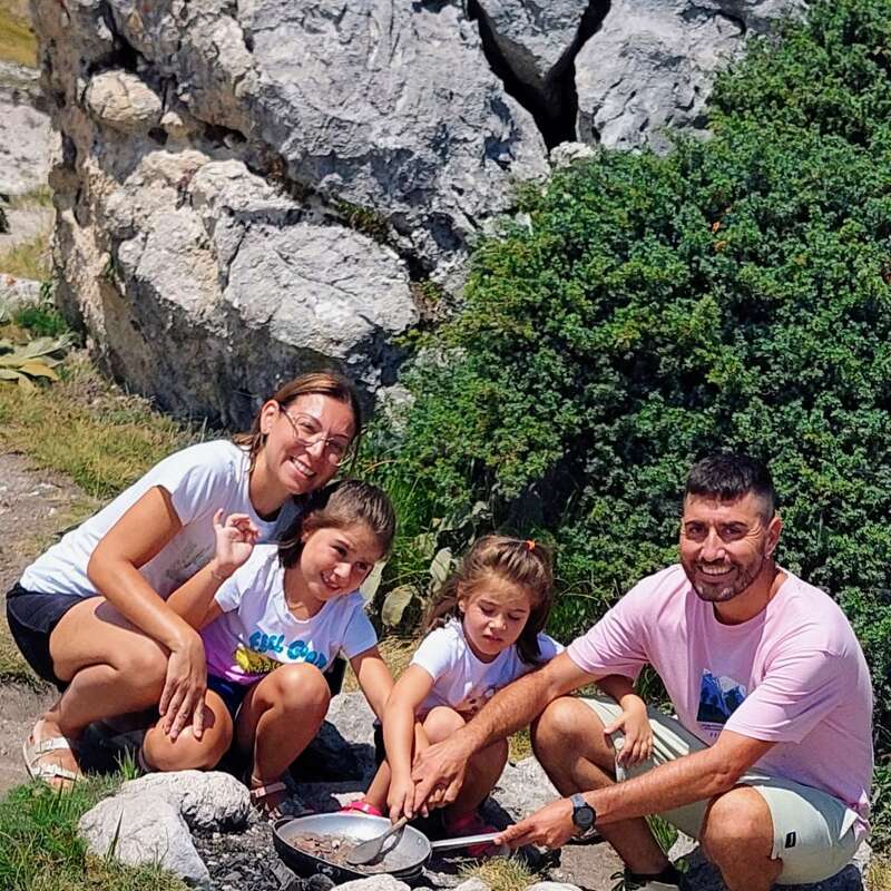 Una familia de cuatro miembros disfruta de una comida al aire libre en una zona rocosa y montañosa. Sonrientes, se sientan juntos alrededor de una hoguera, rodeados de naturaleza y bajo un sol radiante.