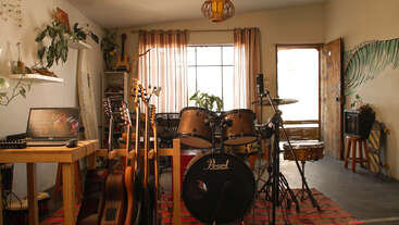 A cozy music studio with guitars, a drum set, laptop on a wooden table, houseplants, and soft natural light filtering through large windows and sheer curtains.
