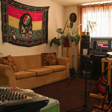 A cozy home recording studio with a tan couch, Bob Marley tapestry, music equipment, guitars, plants, and a bright window with sheer curtains. Creative atmosphere.