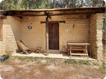 A rustic adobe cabin with a wooden door and simple porch. Wooden chairs, a table, and potted plants add charm. Surrounded by forest and sunlight.