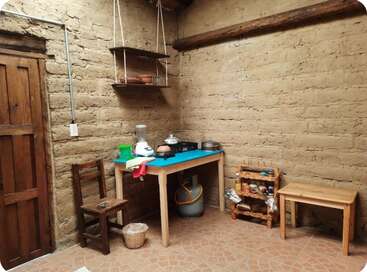 This is a rustic kitchen with adobe walls, a wooden door, simple furniture, basic appliances, shelves, a gas cylinder, utensils, and traditional kitchenware.