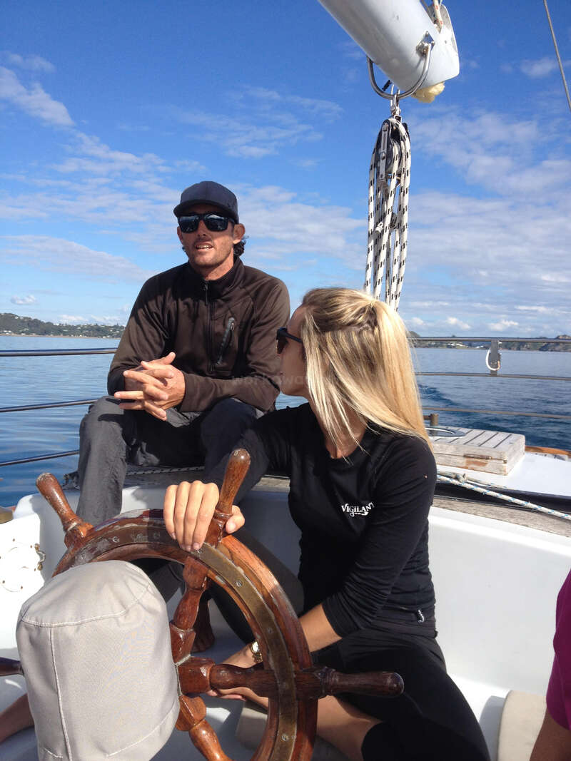 Dos personas están sentadas en un velero. La mujer gobierna con un timón de madera, mientras el hombre está sentado a su lado. Ambos llevan gafas de sol, disfrutando del tiempo soleado.