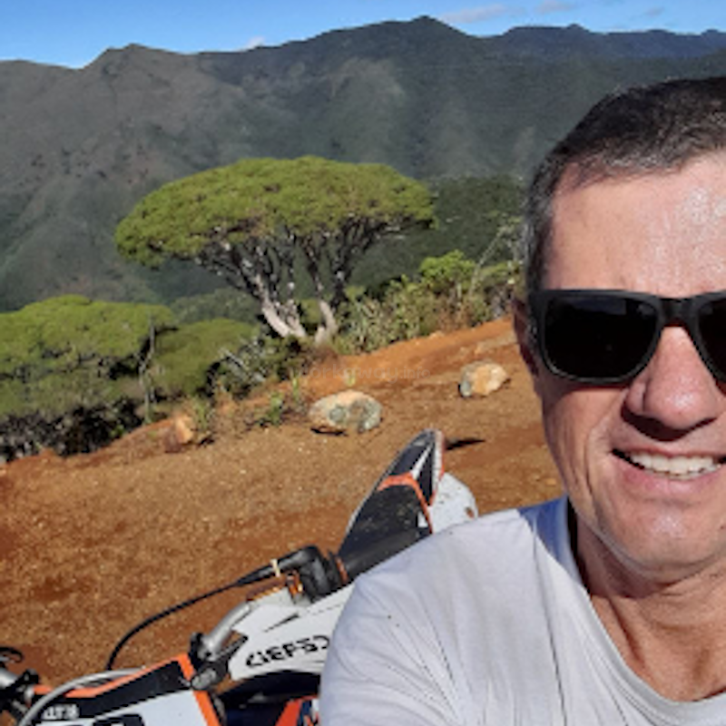 Das Bild zeigt einen Mann mit Sonnenbrille, der ein Selfie mit einem Motorrad vor einer bergigen Landschaft mit Bäumen und Felsen macht.