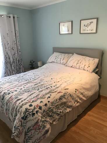 La imagen muestra un dormitorio sereno con una cama con edredón y almohadas florales, un cabecero gris y una pared azul claro adornada con dos cuadros enmarcados.