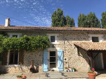 La imagen representa una encantadora casa de piedra con tejado de tejas, contraventanas azules y una chimenea, rodeada de exuberante vegetación y un sereno cielo azul.
