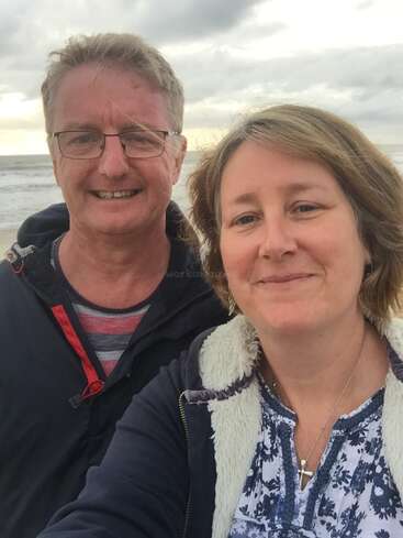 La imagen muestra un selfie de un hombre y una mujer de pie en una playa, con el océano y el cielo nublado visibles detrás de ellos. Van vestidos con ropa informal.