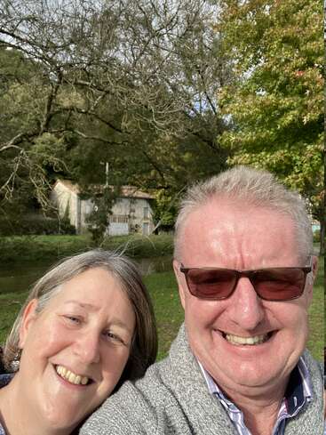 La imagen muestra un selfie de un hombre y una mujer delante de una casa y árboles, con la mujer a la izquierda y el hombre a la derecha, ambos sonriendo.