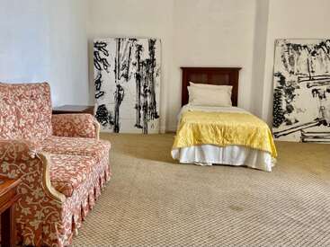 Cette chambre comprend un lit simple avec une couverture jaune, des œuvres d'art abstraites en noir et blanc, un canapé à motifs, une moquette beige et des murs blancs simples et minimaux.
