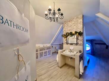 Cette salle de bains mansardée confortable présente des murs en pierre rustique, des planchers en bois, un lustre, un miroir décoratif, un lavabo, une guirlande florale, un panneau étiqueté et une fenêtre en vitrail avec une lumière bleue.