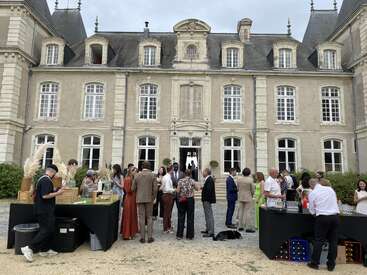 Un groupe de personnes bien habillées se mêle à la foule et socialise à l'extérieur d'un grand bâtiment historique. Des tables avec des boissons et de la nourriture sont installées pour la réunion en plein air.