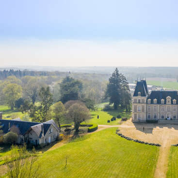 Un grand château se dresse au milieu de vastes pelouses verdoyantes et de jardins bien entretenus. Les arbres environnants et les champs lointains créent un paysage de campagne serein et pittoresque sous un ciel dégagé.