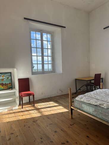 A simple, sunlit room features a wooden floor, single bed with mattress, red chairs, desk, shelving, and large window offering a view of blue sky.