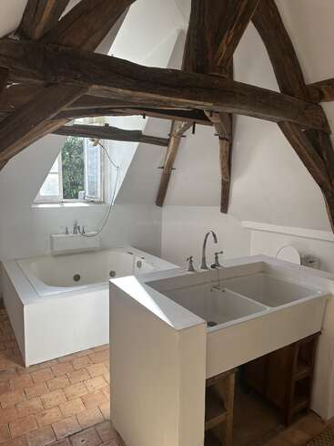 This image shows a rustic bathroom with exposed wooden beams, a large white Jacuzzi tub, double sink, terracotta tile floor, and a window letting in sunlight.