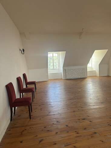 This image shows a spacious, sunlit room with wooden floors, three red chairs against a white wall, slanted ceilings, two windows, and a radiator.