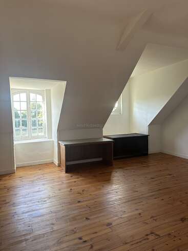 This is a bright, empty room with wooden floors, white walls, slanted ceiling, two desks, and large windows letting in plenty of natural light.