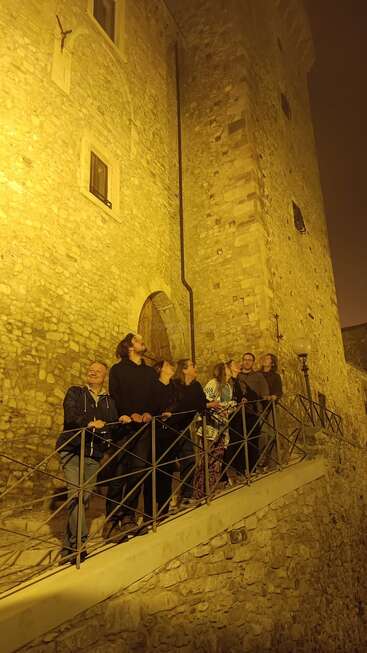 A group of people stand on a stone walkway at night, gazing upward, illuminated by yellow lights against the backdrop of an ancient stone building.