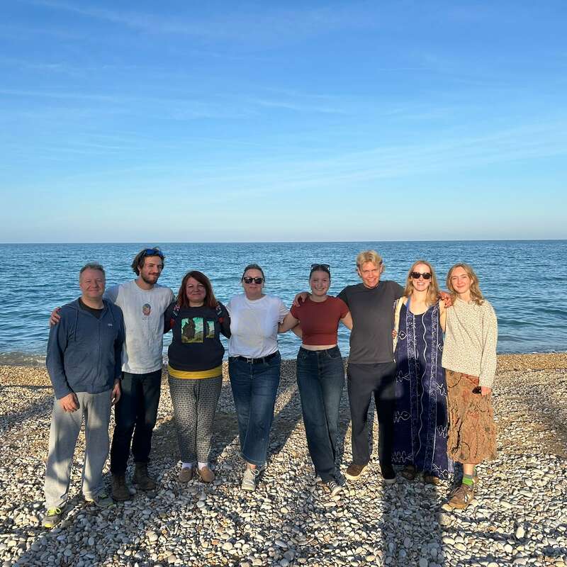 Eight people stand side by side on a rocky beach, smiling at the camera. The sea and a clear blue sky are in the background.