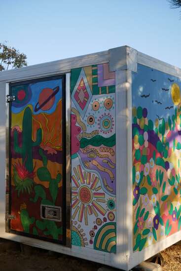 A brightly painted box displays colorful murals of cacti, abstract patterns, sunshine, planets, birds, and flowers, creating a lively and cheerful outdoor artwork scene.